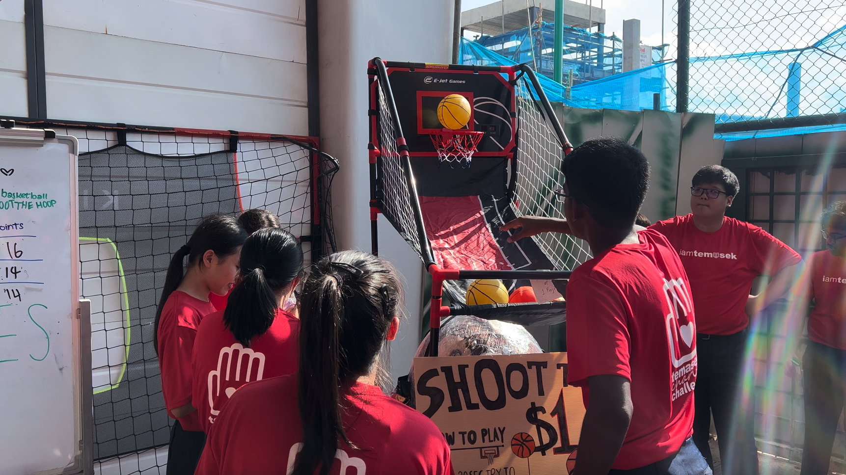 People in red Temasek shirts gather around an arcade basketball hoop game with a cardboard sign reading "Shoot $1."